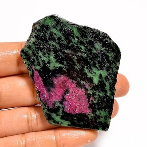 Pode incluir: Uma gema bruta com um padrão marcante de preto, verde e magenta. A superfície da pedra tem uma aparência texturizada, com a cor magenta concentrada no centro. A forma geral é irregular, mostrando sua forma natural.