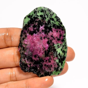Pode incluir: Uma gema em bruto com uma paleta de cores vibrante. A pedra apresenta uma combinação marcante de magenta profundo, preto salpicado e manchas verdes. A forma natural e irregular da pedra aumenta o seu apelo único.