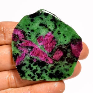 May include: A raw gemstone with a green base, dotted with black specks. Vivid pink-red crystal formations are visible, creating a striking contrast. The stone is held in a hand. The gemstone's irregular shape is visible.