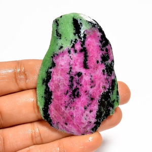 Op de afbeelding: Een ruwe edelsteen met een levendig kleurenpalet. De steen heeft een opvallende combinatie van roze, groene en zwarte tinten. Het roze gebied is het grootst, met zwarte spikkels en een groene rand. De steen wordt in een hand gehouden.