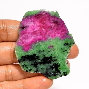 Pode incluir: Uma gema em bruto com uma paleta de cores vibrantes. A seção superior é um magenta profundo, passando para um verde brilhante no meio, com inclusões pretas na parte inferior. A pedra é segurada em uma mão.