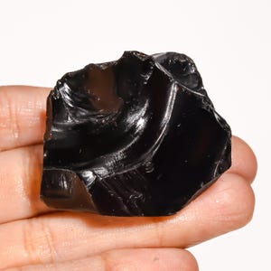 May include: A raw, unpolished piece of black obsidian stone with a smooth, glass-like texture and sharp, irregular edges. The surface of the volcanic rock reflects light, showcasing a deep, solid black color throughout its entire structure.