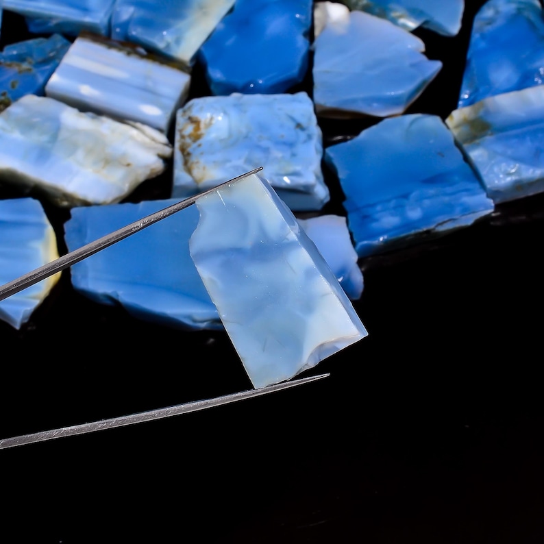 May include: A collection of raw, translucent blue opal stones with irregular shapes and smooth, polished surfaces. One individual piece is held by thin metal tweezers, showcasing its pale blue color, cloudy internal texture, and sharp edges.