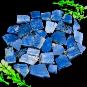May include: A collection of irregular, polished blue opal stones with varying shades of sky blue and white, featuring smooth surfaces and some dark mineral inclusions, arranged in a cluster with bright green plastic foliage accents.