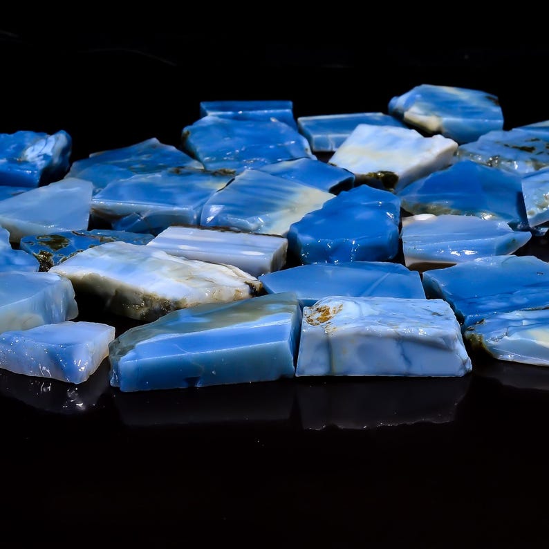 May include: A collection of irregular, raw blue opal fragments with varying shades of sky blue, deep azure, and milky white. Each piece features a smooth, polished surface with natural edges and internal color variations, scattered across a dark field.