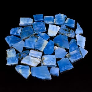 May include: A collection of irregular, flat-cut blue opal stones with varying shades of sky blue and white marbling. Each piece features a smooth, polished surface with unique natural patterns and jagged edges, arranged in a cluster on a black background.