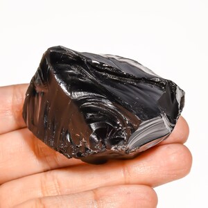 May include: A raw, unpolished piece of black obsidian stone with a glossy, glass-like texture. The mineral features sharp, irregular edges and a deep, reflective black surface with visible conchoidal fractures throughout its structure.