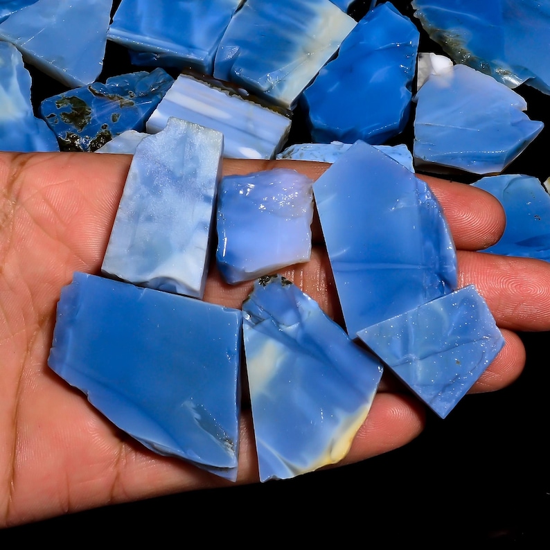 May include: A collection of raw, unpolished blue opal stones with smooth, translucent surfaces and varying shades of sky blue, cerulean, and white. The stones feature irregular, angular shapes with sharp edges and a glossy, vitreous luster throughout.