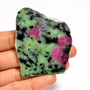 Pode incluir: Uma gema bruta com uma vibrante mistura de cores. A pedra apresenta uma base verde clara salpicada de preto e acentuada por manchas rosa brilhante. Sua forma irregular e textura natural lhe dão um apelo único e orgânico.