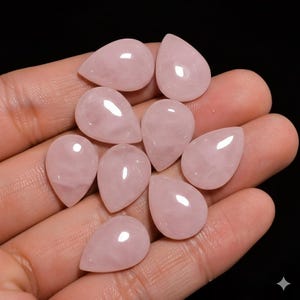 Pode incluir: Uma coleção de nove pedras preciosas de quartzo rosa polidas, cada uma em forma de gota. As pedras têm um tom rosa suave e uma superfície lisa e refletora. As pedras preciosas são exibidas em um fundo escuro.
