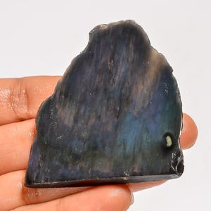 May include: A polished, irregularly shaped gemstone with a dark, iridescent surface. The stone displays a mix of deep blues, purples, and hints of green, with a smooth, reflective quality. The gemstone is held in a hand.