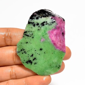 Pode incluir: Uma pedra preciosa em bruto com uma paleta de cores vibrante. A pedra apresenta uma combinação marcante de tons verdes, rosa e pretos. A base verde é salpicada com inclusões pretas, enquanto uma mancha de rosa vivo adiciona um toque de cor. A borda superior é preta.