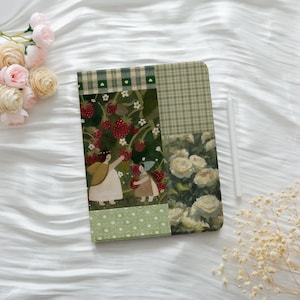 May include: A decorative journal with a patchwork design featuring illustrations of strawberries, white roses, and green plaid patterns. A white stylus rests on the journal's surface, with pink roses and dried flowers in the background.