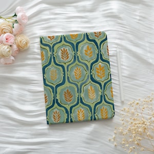 May include: A rectangular journal with a repeating pattern of teal, green, and gold leaf-like shapes. A white stylus rests on the right side. The journal is on a white, textured surface, with pink and cream-colored flowers in the upper left corner.