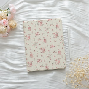 May include: A cream-coloured journal with a floral pattern of pink roses and green leaves. A white stylus rests beside the journal. Pink roses and dried flowers are arranged on a white, textured surface.