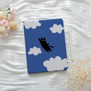 May include: A blue notebook with a whimsical design featuring a black cat soaring through a sky with white clouds. A white stylus rests beside the notebook. The notebook is surrounded by flowers.