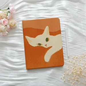 May include: An orange tablet case with a playful design of a white cat with green eyes and a pink nose. A white stylus is placed next to the case. The case is on a white, textured surface, with pink flowers and dried white flowers in the background.