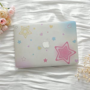 May include: A clear laptop case adorned with pastel stars and hearts. The case features a gradient of colors, including pink, blue, and yellow. A large pink star is prominently displayed, along with the Apple logo.