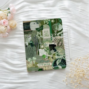 May include: A green and white collage-style journal with various images and text, including butterflies, stars, and the phrases "Good Things Take Time" and "Keep Going." A white stylus rests beside the journal. The background is a white, textured surface.