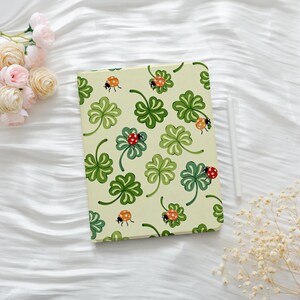 May include: A rectangular tablet case featuring a light green background with a repeating pattern of dark green four-leaf clovers and small red ladybugs with black spots. A white stylus pen is positioned vertically along the right side.
