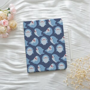 May include: A blue tablet case featuring a repeating pattern of cartoon sharks in light blue and white. The sharks have red mouths and are set against a dark blue background. A white stylus rests beside the case.