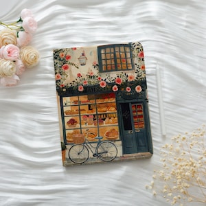 May include: A notebook with a watercolor illustration of a French patisserie. The image features a bakery with a blue door, window, and a bicycle. The facade is adorned with flowers and the word "Patisserie".