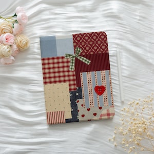 May include: A patchwork journal with a variety of fabric patterns, including red plaid, blue stripes, and floral prints. A green and white checkered bow is tied on the cover, and a red heart-shaped button is attached. A white stylus rests beside the journal.