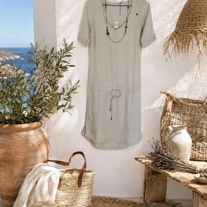 May include: A light sage green short-sleeved dress featuring a V-neckline, a drawstring waist with thin ties, and a small brown button detail on the left shoulder. A long, delicate necklace with dark beads hangs around the neckline of the dress.