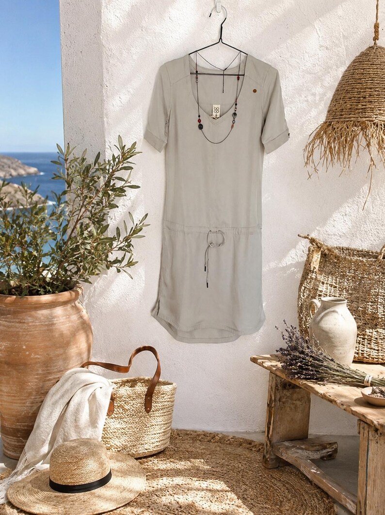 May include: A light sage green short-sleeved dress featuring a V-neckline, a drawstring waist with thin ties, and a small brown button detail on the left shoulder. A long, delicate necklace with dark beads hangs around the neckline of the dress.