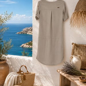 May include: A light beige knee-length dress with short sleeves, a round neckline, and a small brown rectangular brand tag on the upper back. The dress features a subtle vertical pleat at the center back and rolled sleeve cuffs with button tabs.