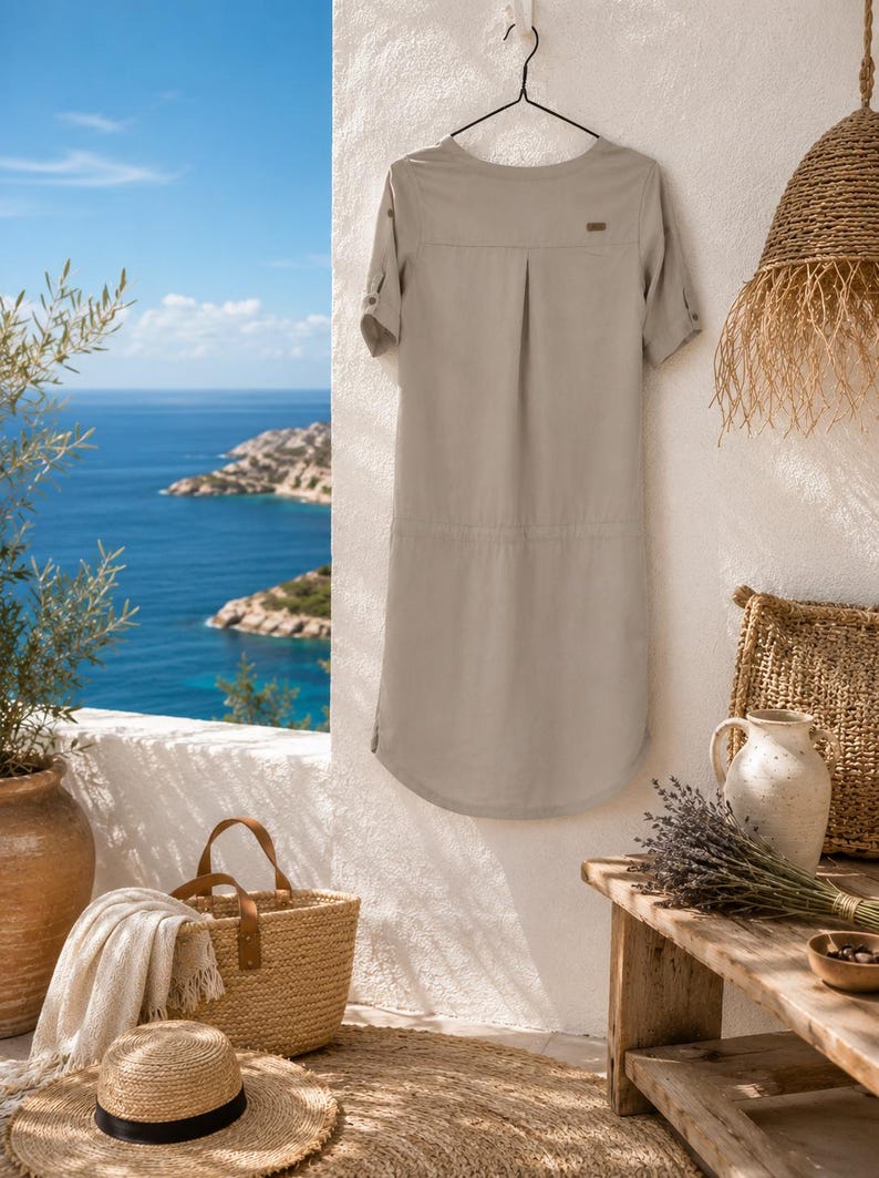 May include: A light beige knee-length dress with short sleeves, a round neckline, and a small brown rectangular brand tag on the upper back. The dress features a subtle vertical pleat at the center back and rolled sleeve cuffs with button tabs.