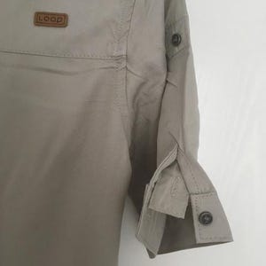 May include: A light beige button-down shirt featuring a small brown rectangular patch on the chest with the text "LOOP" embossed in white. The shirt has three-quarter length sleeves with buttoned cuffs and a tab detail on the upper arm.