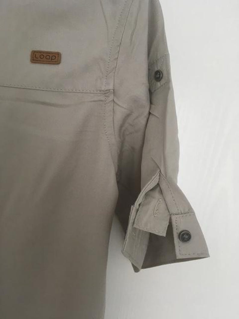 May include: A light beige button-down shirt featuring a small brown rectangular patch on the chest with the text "LOOP" embossed in white. The shirt has three-quarter length sleeves with buttoned cuffs and a tab detail on the upper arm.
