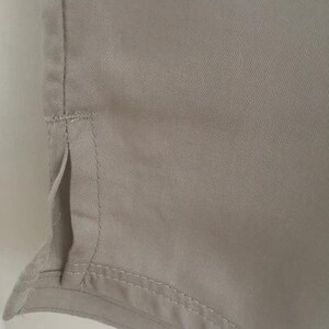 May include: A light beige garment featuring a smooth, woven fabric texture with a vertical side seam detail. The hem includes a small, open side vent with reinforced stitching along the edges, showcasing a clean, finished construction throughout.