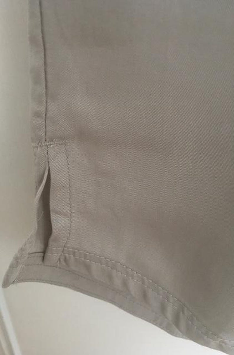 May include: A light beige garment featuring a smooth, woven fabric texture with a vertical side seam detail. The hem includes a small, open side vent with reinforced stitching along the edges, showcasing a clean, finished construction throughout.