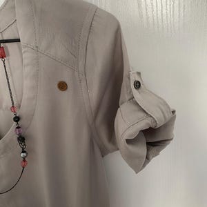 May include: A light beige button-down shirt with a V-neckline, featuring a small circular wooden logo patch on the chest and sleeves that are rolled up and secured with a button tab. A beaded necklace with pink, black, and silver beads is visible.