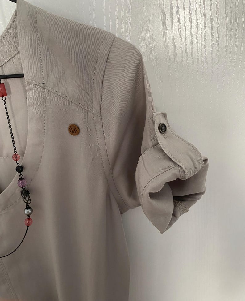 May include: A light beige button-down shirt with a V-neckline, featuring a small circular wooden logo patch on the chest and sleeves that are rolled up and secured with a button tab. A beaded necklace with pink, black, and silver beads is visible.
