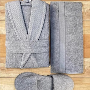 Può includere: Un set spa grigio composto da un accappatoio in spugna, un asciugamano piegato e un paio di pantofole abbinate. L'accappatoio ha un collo a scialle e una cintura in vita. L'asciugamano ha una striscia decorativa. Gli articoli sono disposti su una superficie di legno.