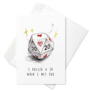 May include: A white greeting card with a D20 die illustration. The die is gray with red hearts and the number 20 on two sides. The text "I rolled a 20 when I met you" is below the die. The card is inside a white envelope.