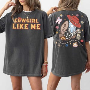 May include: A charcoal gray short-sleeve t-shirt featuring the text COWGIRL LIKE ME in yellow and red on the front. The back displays a graphic of a cow-print cowboy boot, a red hat, a pink bow, and blocks with the letters T, S, and 13.