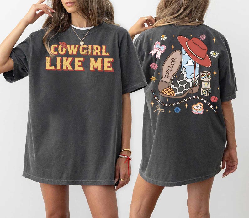 May include: A charcoal gray short-sleeve t-shirt featuring the text COWGIRL LIKE ME in yellow and red on the front. The back displays a graphic of a cow-print cowboy boot, a red hat, a pink bow, and blocks with the letters T, S, and 13.