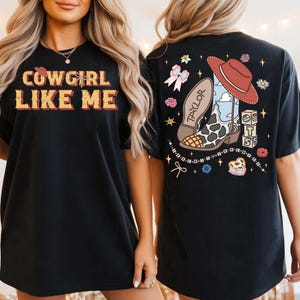 May include: A black t-shirt featuring the text COWGIRL LIKE ME on the front. The back displays a graphic of a cowboy boot with a cow print pattern, a red hat, the name TAYLOR, the number 13, and stacked blocks with the letters T, S, and B.