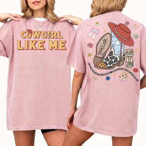 May include: A dusty rose t-shirt featuring the text COWGIRL LIKE ME on the front. The back displays a graphic of a cow-print cowboy boot, a red hat, blocks with the letters T S, and a beaded string with the text I KNEW IT WHEN I KNEW YOU.