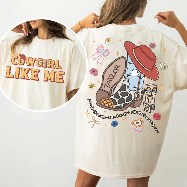 May include: An off-white t-shirt featuring a graphic print. The front displays the text "COWGIRL LIKE ME" in orange and red. The back shows a cow-print boot, a red hat, blocks with "T S", and a beaded string with the text "I KNEW IT WHEN I MET YOU".