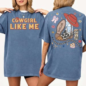 May include: A blue t-shirt featuring the text COWGIRL LIKE ME on the front. The back displays a graphic of a cow-print cowboy boot, a red hat, a bow, and blocks with the letters T S, surrounded by stars and flowers with the text TAYLOR.