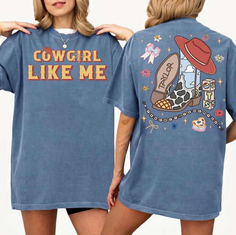 May include: A blue t-shirt featuring the text COWGIRL LIKE ME on the front. The back displays a graphic of a cow-print cowboy boot, a red hat, a bow, and blocks with the letters T S, surrounded by stars and flowers with the text TAYLOR.