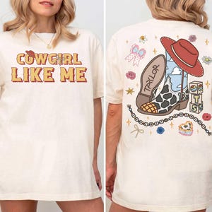 May include: An off-white t-shirt featuring the text COWGIRL LIKE ME on the front. The back displays a graphic of a cow-print cowboy boot, a red hat, blocks with the letters T S, and a beaded string with the text THE ERAS TOUR.