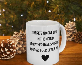 May include: White ceramic mug with a black heart and the text: "THERE'S NO ONE ELSE IN THE WORLD I'D RATHER HAVE SNORING LOUD AS FUCK BESIDE ME." The mug is on a wooden surface with pine cones in the background.