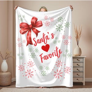 May include: White blanket with a large red bow, snowflakes, and the text "Santa's Favorite" in red. Green and red snowflakes are scattered across the blanket. A festive holiday-themed throw blanket.