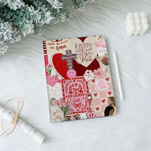 Peut inclure: Un journal décoratif avec un design de collage comprenant des cœurs, des fleurs et des phrases inspirantes en plusieurs langues. La couverture comprend un cœur rouge, une croix et les mots "You Deserve Joy". Un stylet blanc est posé à côté.